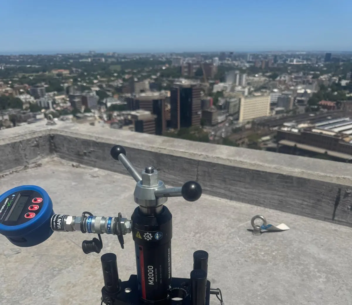 Hydraulic load tester on a high-rise roof proof testing a roof anchor, with the Sydney skyline behind